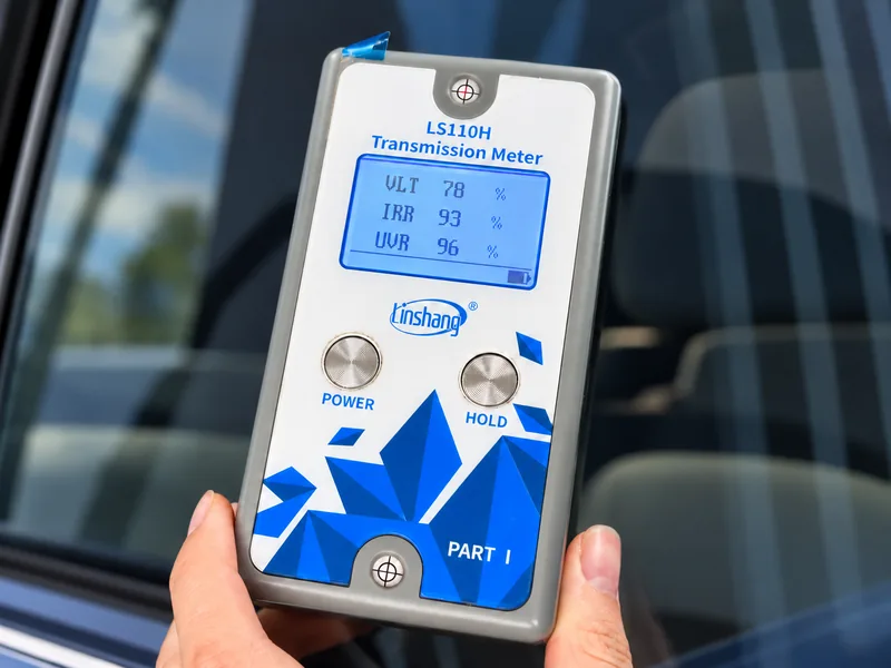 Handheld transmission meter displaying illustrative VLT, IRR and UVR readings beside automotive glass.